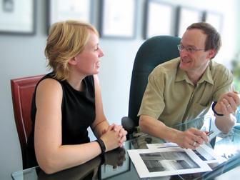 Photograph of a man and woman talking whilst sitting at a desk in a bright office with artwork on the desk in front of them.Blurred artwork is on the walls behind them.