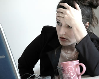 Photograph of a stressed looking business woman holding her forehead, looking at a PC screen, a coffee mug on the desk by her elbow.