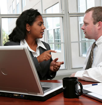 Photograph taken in a modern office of an Indian woman sitting at a desk in front of a laptop gesturing and talking to a caucasian man sitting to her side.