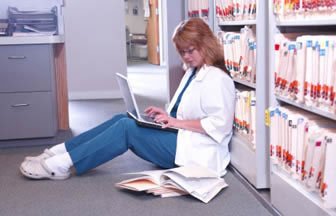 Photograph a mdeical professional sitting on the floor leaning against a wall of medical files, reading a book from the floor on top of a pile of papers whislt she types on a laptop which is balanced on her knees. 