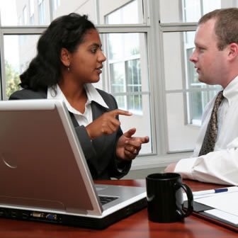 Photograph taken in a modern office of an Indian woman sitting at a desk in front of a laptop gesturing and talking to a caucasian man sitting to her side.