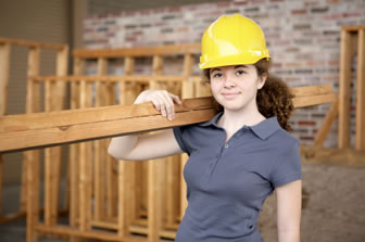 Young woman wearing a navy top and a yellow hard hard carried a plank of wood from a roofing structure over her shoulder.