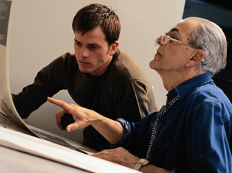 A young man and an older man look at a series of printed photographs on a long stretch of paper.