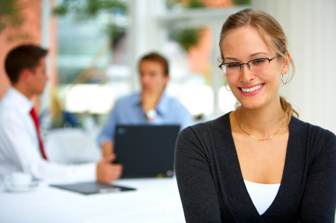 A young woman in business dress is smiling into the camera in the bottom right of the picture, out of focus in the background a couple of men talk, seated at a table in front of a laptop and clipboard.